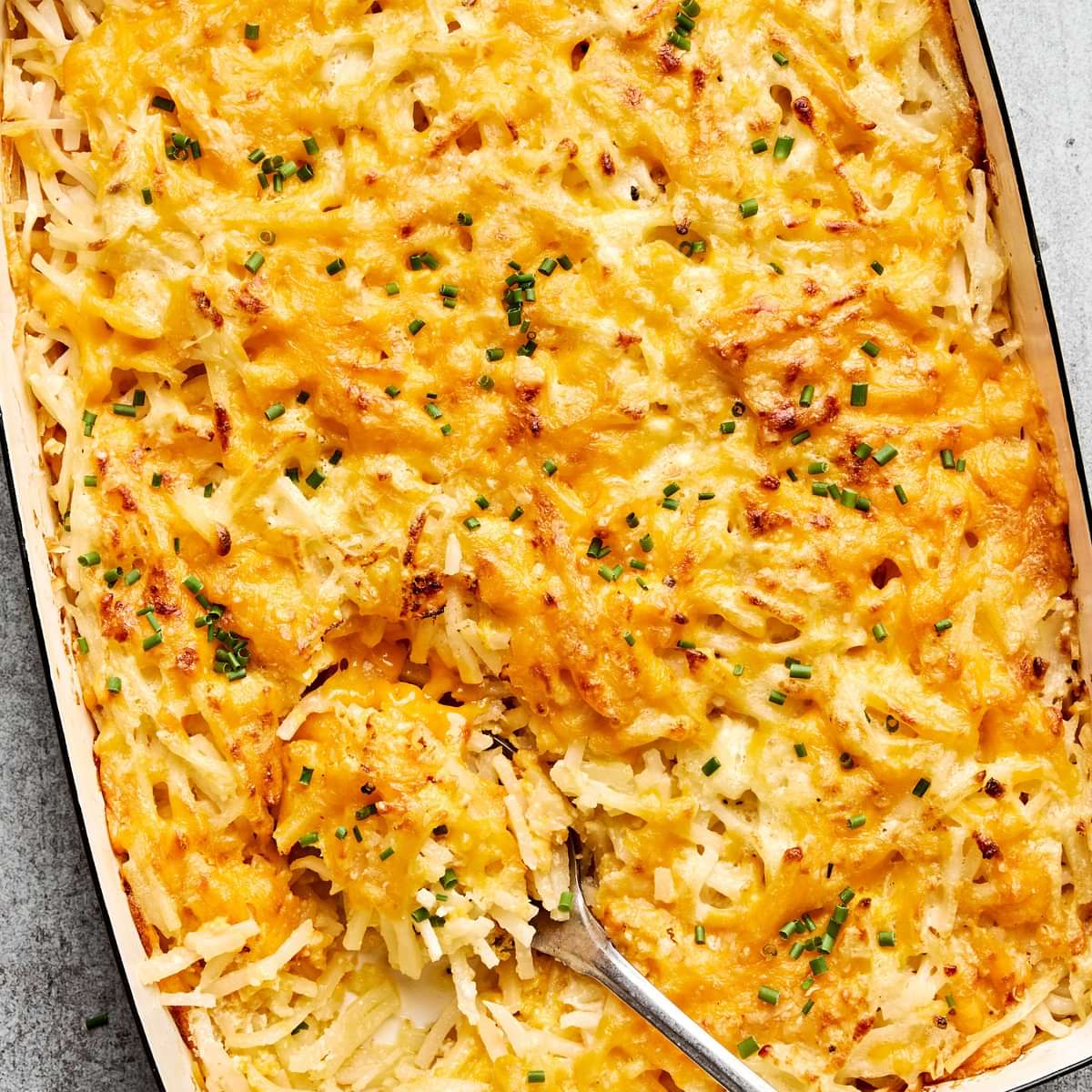 A golden-brown hashbrown casserole in a baking dish, with a creamy potato filling topped with melted cheese and chives