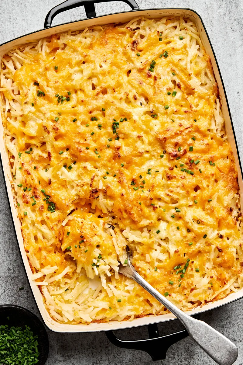 A golden-brown hashbrown casserole in a baking dish, with a creamy potato filling topped with melted cheese and chives