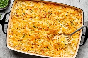 A golden-brown hashbrown casserole in a baking dish, with a creamy potato filling topped with melted cheese and chives