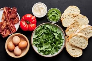 8 bread slices, arugula, 4 eggs, cooked bacon, heirloom tomatoes, mayo and pesto in prep bowls to make a BLT with Pesto