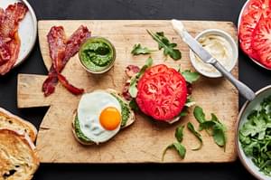a homemade heirloom tomato BLT sandwich with pesto being assembled on a cutting board