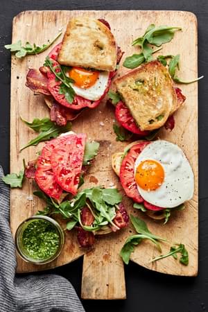 heirloom BLT sandwiches with pesto and fried eggs being assembled on a cutting board