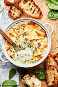 hot spinach and artichoke dip in a baking dish being scooped with a spoon surrounded by toasted crostini for serving