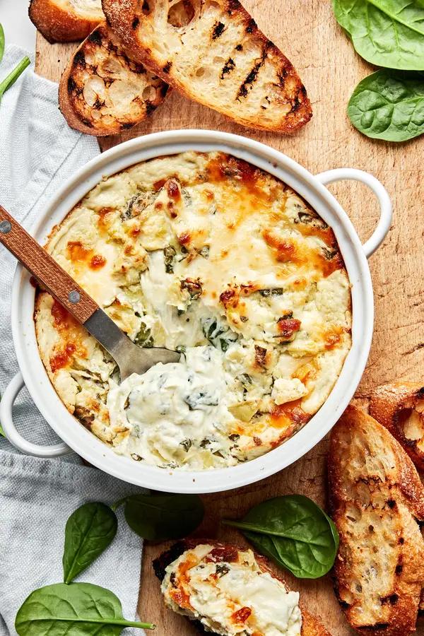 hot spinach and artichoke dip in a baking dish being scooped with a spoon surrounded by toasted crostini for serving