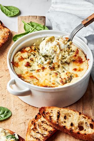 hot spinach and artichoke dip in a baking dish being scooped with a spoon surrounded by toasted crostini for serving