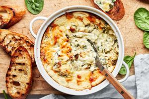 hot spinach and artichoke dip in a baking dish being scooped with a spoon surrounded by toasted crostini for serving