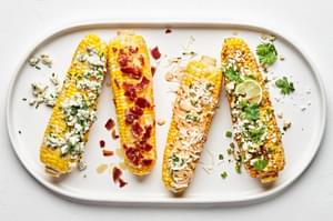 4 grilled corn on the cob with different toppings: maple bourbon butter, chipotle mayo, spicy cotija cheese, & blue cheese