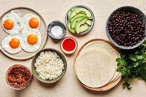tortillas, black beans, fried eggs, avocado, salsa, cilantro and cotija cheese in prep bowls to make huevos rancheros