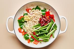 bell peppers, green onions, peanuts, ginger, garlic, and chili peppers being cooked in sesame oil in a skillet