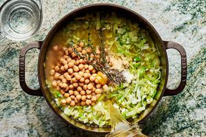 jar pouring chicken stock into a pot of cooked leeks, onions and garlic with garbanzo beans and fresh thyme sprigs.