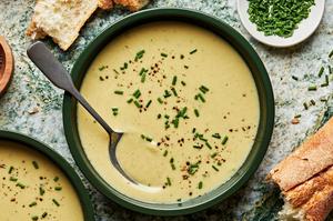 a spoon dipping into a green bowl filled with leek soup topped with fresh chives and surrounded by crusty bread and chives.
