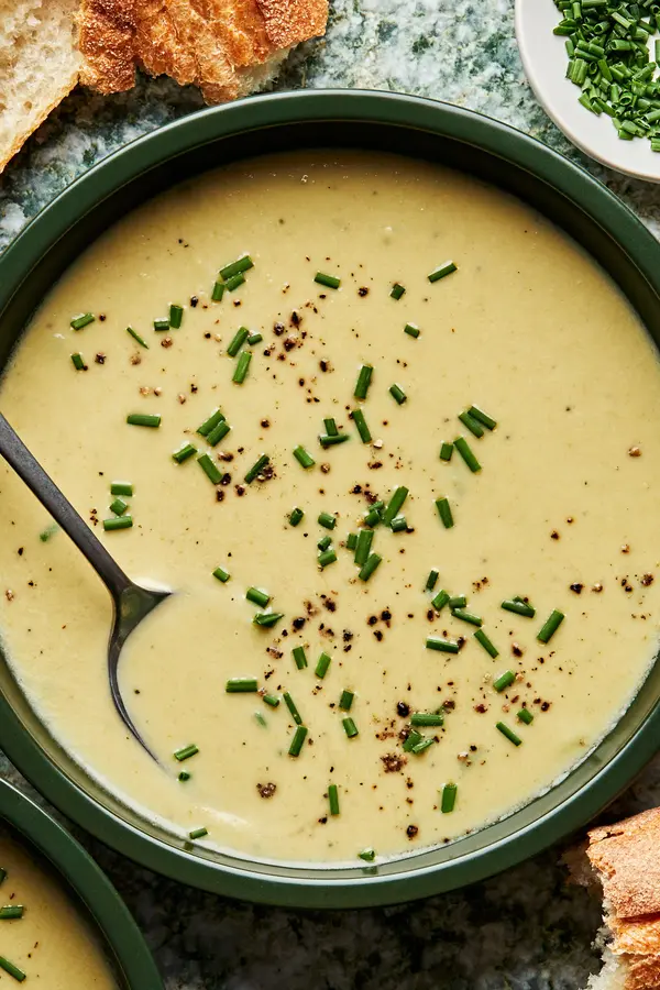 Bowl of creamy leek soup topped with fresh chives and a spoon in it surrounded by crusty bread slices.