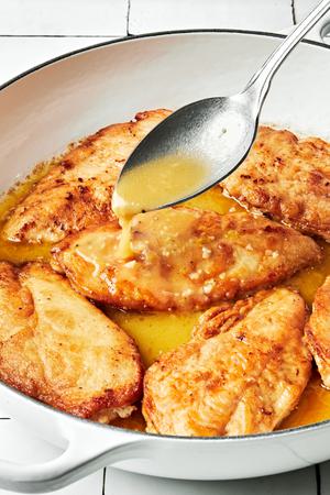 homemade lemon sauce being spooned over lightly breaded chicken in a pan