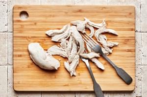 cooked chicken breast being shredded with two forks on a cutting board