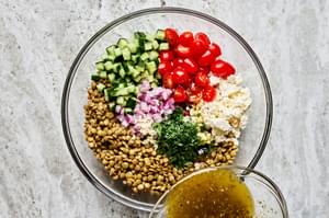 dressing being poured over lentils, cucumber, tomatoes, onion, parsley, lemon juice & feta cheese in a glass bowl