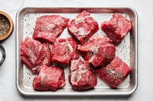 raw chuck roast on a sheet pan, cut into 3 inch pieces being seasoned with salt and pepper