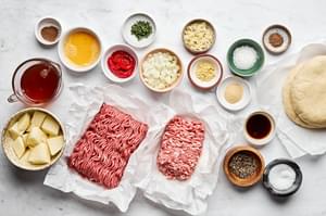 Meat pie ingredients laid out, including pork, beef, onion, herbs, spices and a pie crust ready for assembly.