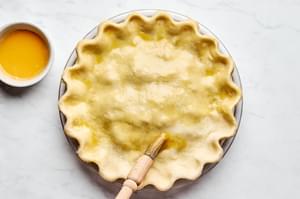 an unbaked meat pie filled with potatoes, ground beef and ground pork being brushed with egg wash to prepare to bake