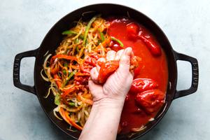 Hand crushing canned tomatoes into a skillet with onions, bell peppers and spices for meatball shakshuka