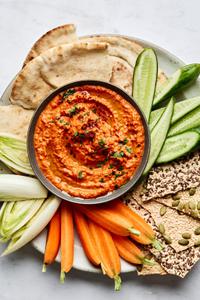 a bowl of homemade Muhammara dip surrounded by crackers, carrots, cucumber and pita for dipping