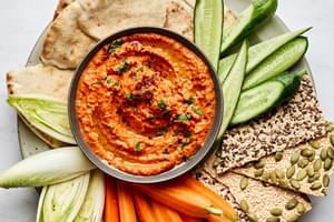 a bowl of homemade Muhammara dip surrounded by crackers, carrots, cucumber and pita for dipping
