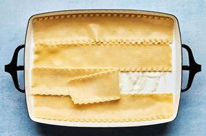 homemade white sauce spread in the bottom of a casserole dish being topped with cooked lasagna noodles