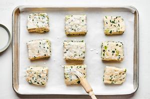 onion chive biscuit dough cut into 9 pieces on a parchment lined baking sheet being brushed with buttermilk