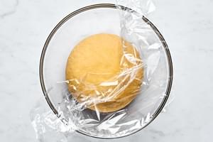 homemade kneaded pasta dough in a glass bowl covered with plastic wrap