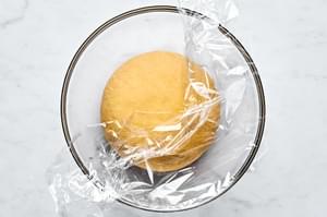 homemade kneaded pasta dough in a glass bowl covered with plastic wrap