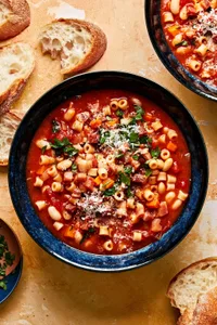 a bowl of homemade pasta e fagioli sprinkled with parsley and parmesan surrounded by slices of crusty bread