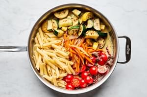 pasta, zucchini, squash, sun dried tomatoes, cherry tomatoes and sauce being tossed in a skillet