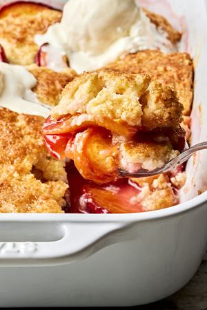 homemade peach cobbler topped with vanilla ice cream being scooped out of a baking dish