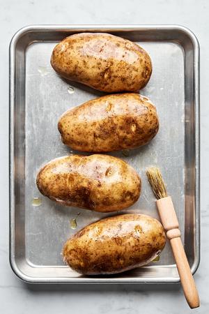 4 russet potatoes being brushed with olive oil on a rimmed baking sheet