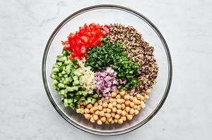 quinoa, chickpeas, red onion, cucumber, tomatoes, garlic, parsley and jalapeños being tossed in a bowl