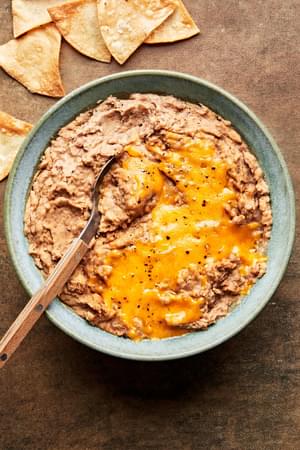 homemade refried beans topped with melted cheddar cheese in a bowl with a spoon surrounded by tortilla chips