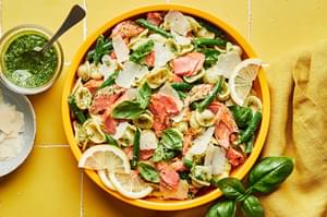 salmon pasta with homemade pesto in a serving bowl next to a jar of pesto and a bowl of shaved parmesan for topping