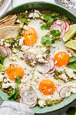 salsa verde baked eggs in a skillet sprinkled with cotija, cilantro and radishes being served with corn tortillas and limes