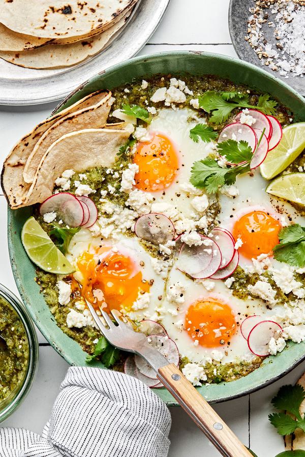 salsa verde baked eggs in a skillet sprinkled with cotija, cilantro and radishes being served with corn tortillas and limes