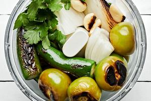roasted tomatillos, onions and jalapeños being placed in a food processor with  cilantro and salt