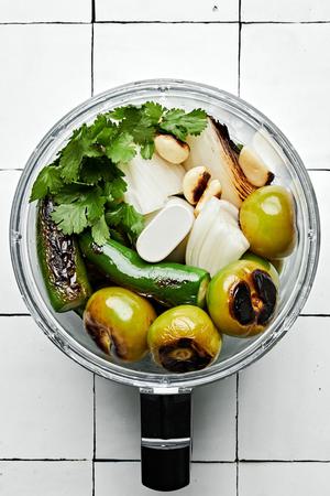 roasted tomatillos, onions and jalapeños being placed in a food processor with cilantro and salt