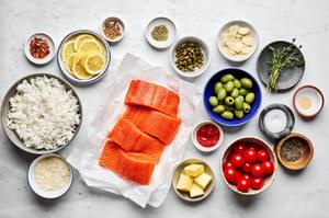 4 salmon filets, butter, tomato paste, spices, lemon, tomatoes, olives, and capers in bowls to make salmon en papillote