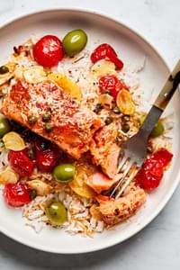 Cooked salmon, on a plate served over rice with a tomato butter sauce toped with tomatoes, green olives & capers