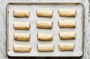 twelve uncooked sausage rolls arranged on a rimmed baking sheet and brushed with egg