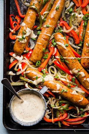 Italian sausage heroes on a rimmed baking sheet with sliced onions and bell peppers with a bowl of honey mustard sauce