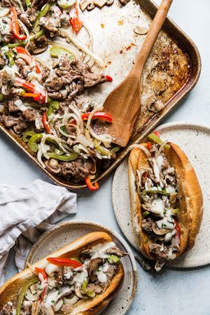 sheet pan philly cheesesteaks being scooped from a baking sheet onto toasted hoagie rolls