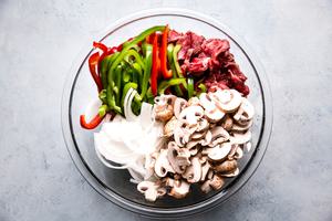 onions, bell peppers, thinly sliced steak and mushrooms in a glass bowl for homemade Philly cheese steaks