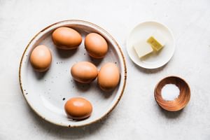 6 eggs on a plate next to 2 tablespoons of butter and salt in a small wooden bowl
