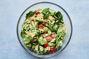 spinach, cooked orzo, tomatoes, feta and basil vinaigrette being tossed in a glass bowl