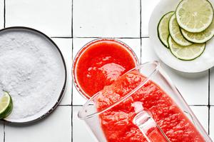a blended strawberry margarita being poured in a slat rimmed glass next to lime slices for garnish