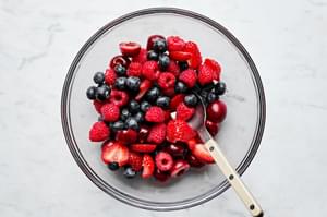 fresh strawberries, blueberries, raspberries and chopped cherries being stirred together in a bowl